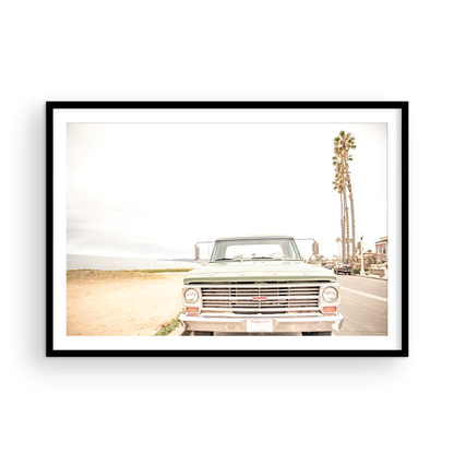 Nostalgic Southern California beach art featuring a classic light green Ford F-Series pickup truck parked by the San Diego coast. This horizontal surf print captures the vintage vehicle with a backdrop of iconic tall palm trees, a hazy beach, and the ocean, embodying surf vibes and perfect for coastal farmhouse decor. Photograph in minimalist framed mock up.