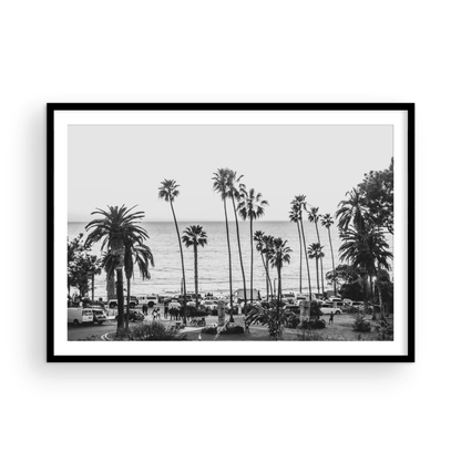 A classic black and white horizontal print of Swamis, the iconic San Diego surf spot, showcasing a panoramic view from above. This piece of Southern California travel photography features dozens of silhouetted palm trees against a bright sky, with a lively beach scene of cars and people below, stretching towards the ocean. Perfect as timeless beach art or neutral coastal decor for a surf vibe. Photograph in minimalist framed mock up.