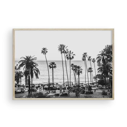 A classic black and white horizontal print of Swamis, the iconic San Diego surf spot, showcasing a panoramic view from above. This piece of Southern California travel photography features dozens of silhouetted palm trees against a bright sky, with a lively beach scene of cars and people below, stretching towards the ocean. Perfect as timeless beach art or neutral coastal decor for a surf vibe. Photograph in minimalist framed mock up.