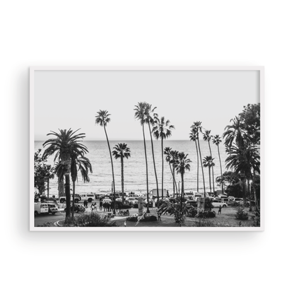 A classic black and white horizontal print of Swamis, the iconic San Diego surf spot, showcasing a panoramic view from above. This piece of Southern California travel photography features dozens of silhouetted palm trees against a bright sky, with a lively beach scene of cars and people below, stretching towards the ocean. Perfect as timeless beach art or neutral coastal decor for a surf vibe. Photograph in minimalist framed mock up.