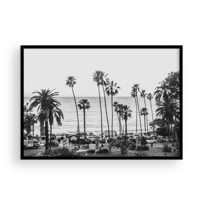 A classic black and white horizontal print of Swamis, the iconic San Diego surf spot, showcasing a panoramic view from above. This piece of Southern California travel photography features dozens of silhouetted palm trees against a bright sky, with a lively beach scene of cars and people below, stretching towards the ocean. Perfect as timeless beach art or neutral coastal decor for a surf vibe. Photograph in minimalist framed mock up.