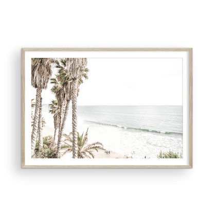 Dreamy coastal palm tree print featuring several tall palm trees with shaggy trunks, and surfers walking down the tranquil beach. This vertical tropical print is captured at Swamis Beach on the Southern California coast, is bathed in bright, hazy sunlight, creating a pastel beach art effect. Evokes serene summer vibes, perfect for beach house decor. Photograph in minimalist surf art mock up.