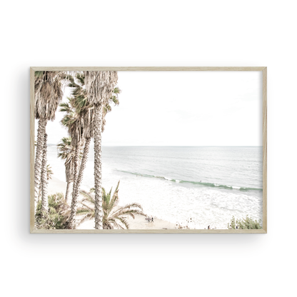 Dreamy coastal palm tree print featuring several tall palm trees with shaggy trunks, and surfers walking down the tranquil beach. This vertical tropical print is captured at Swamis Beach on the Southern California coast, is bathed in bright, hazy sunlight, creating a pastel beach art effect. Evokes serene summer vibes, perfect for beach house decor. Photograph in minimalist mock up.