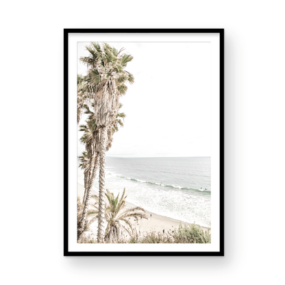 Dreamy coastal palm tree print featuring several tall palm trees with shaggy trunks, lining a gentle slope down to a tranquil beach. This vertical tropical print is captured at Swamis Beach on the Southern California coast, is bathed in bright, hazy sunlight, creating a pastel beach art effect. Evokes serene summer vibes, perfect for beach house decor. Photograph in minimalist surf art mock up.