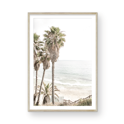 Dreamy coastal palm tree print featuring several tall palm trees with shaggy trunks, next to a staircase down to a tranquil beach. This vertical tropical print is captured at Swamis Beach on the Southern California coast, is bathed in bright, hazy sunlight, creating a pastel beach art effect. Evokes serene summer vibes, perfect for beach house decor. Photograph in minimalist surf art mock up.