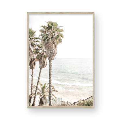 Dreamy coastal palm tree print featuring several tall palm trees with shaggy trunks, next to a staircase down to a tranquil beach. This vertical tropical print is captured at Swamis Beach on the Southern California coast, is bathed in bright, hazy sunlight, creating a pastel beach art effect. Evokes serene summer vibes, perfect for beach house decor. Photograph in minimalist surf art mock up.