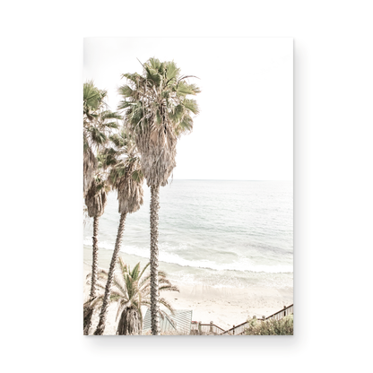 Dreamy coastal palm tree print featuring several tall palm trees with shaggy trunks, next to a staircase down to a tranquil beach. This vertical tropical print is captured at Swamis Beach on the Southern California coast, is bathed in bright, hazy sunlight, creating a pastel beach art effect. Evokes serene summer vibes, perfect for beach house decor. Photograph in minimalist surf art mock up.