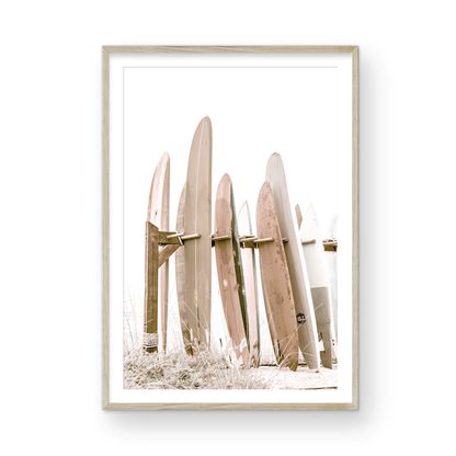 Neutral surfboard wall art. framed fine art photography for the modern coastal home. Minimalist vertical surf art print showcasing a line-up of surfboards in various lengths and soft, neutral pastel hues, resting on a simple wooden rack with some dry grass in the foreground. This piece of surf wall decor features a bright, hazy white backdrop, enhancing its clean aesthetic. Perfect as pastel surf decor or for adding surf vibes to kids surf room decor or a beach house. 