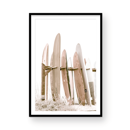 Neutral surfboard wall art. framed fine art photography for the modern coastal home. Minimalist vertical surf art print showcasing a line-up of surfboards in various lengths and soft, neutral pastel hues, resting on a simple wooden rack with some dry grass in the foreground. This piece of surf wall decor features a bright, hazy white backdrop, enhancing its clean aesthetic. Perfect as pastel surf decor or for adding surf vibes to kids surf room decor or a beach house. 