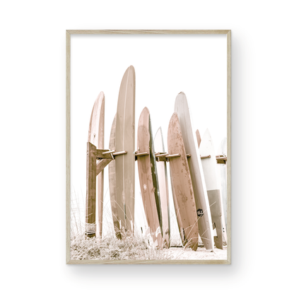 Neutral surfboard wall art. framed fine art photography for the modern coastal home. Minimalist vertical surf art print showcasing a line-up of surfboards in various lengths and soft, neutral pastel hues, resting on a simple wooden rack with some dry grass in the foreground. This piece of surf wall decor features a bright, hazy white backdrop, enhancing its clean aesthetic. Perfect as pastel surf decor or for adding surf vibes to kids surf room decor or a beach house. 