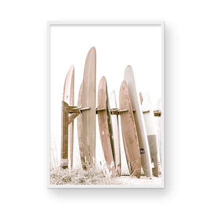 Neutral surfboard wall art. framed fine art photography for the modern coastal home. Minimalist vertical surf art print showcasing a line-up of surfboards in various lengths and soft, neutral pastel hues, resting on a simple wooden rack with some dry grass in the foreground. This piece of surf wall decor features a bright, hazy white backdrop, enhancing its clean aesthetic. Perfect as pastel surf decor or for adding surf vibes to kids surf room decor or a beach house. 