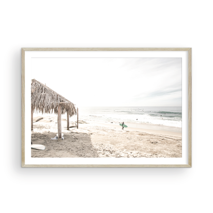 Fine art of WindanSea Beach, La Jolla California horizontal landscape print by Southern California photographer Kim Dybczak of Salt Creek Prints, featuring a bright, slightly desaturated image shows Windansea Beach in La Jolla, California. A thatched beach hut stands on the sandy shore in the left foreground. Further down the beach, a surfer carrying a teal surfboard walks towards the ocean where several other surfers are visible in the water. Photograph in minimalist framed mock up