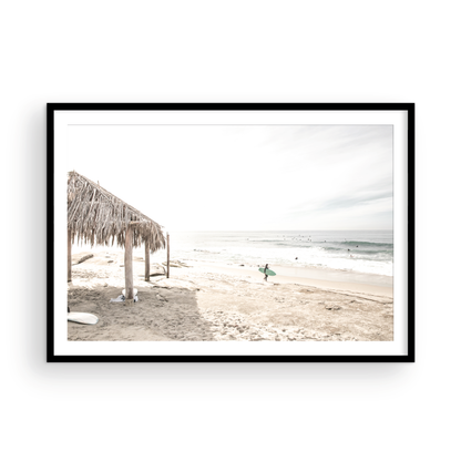 Fine art of WindanSea Beach, La Jolla California horizontal landscape print by Southern California photographer Kim Dybczak of Salt Creek Prints, featuring a bright, slightly desaturated image shows Windansea Beach in La Jolla, California. A thatched beach hut stands on the sandy shore in the left foreground. Further down the beach, a surfer carrying a teal surfboard walks towards the ocean where several other surfers are visible in the water. Photograph in minimalist framed mock up