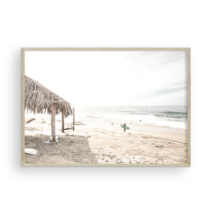 Fine art of WindanSea Beach, La Jolla California horizontal landscape print by Southern California photographer Kim Dybczak of Salt Creek Prints, featuring a bright, slightly desaturated image shows Windansea Beach in La Jolla, California. A thatched beach hut stands on the sandy shore in the left foreground. Further down the beach, a surfer carrying a teal surfboard walks towards the ocean where several other surfers are visible in the water. Photograph in minimalist framed mock up