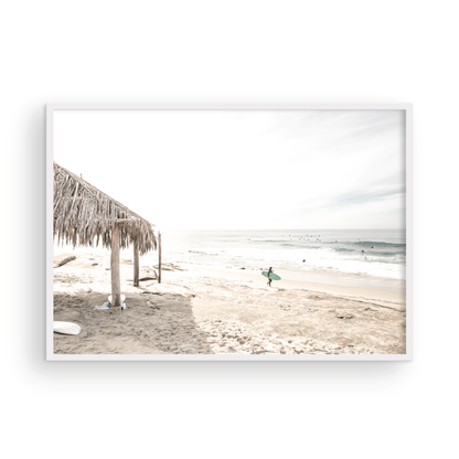 Fine art of WindanSea Beach, La Jolla California horizontal landscape print by Southern California photographer Kim Dybczak of Salt Creek Prints, featuring a bright, slightly desaturated image shows Windansea Beach in La Jolla, California. A thatched beach hut stands on the sandy shore in the left foreground. Further down the beach, a surfer carrying a teal surfboard walks towards the ocean where several other surfers are visible in the water. Photograph in minimalist framed mock up