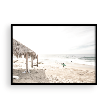 Fine art of WindanSea Beach, La Jolla California horizontal landscape print by Southern California photographer Kim Dybczak of Salt Creek Prints, featuring a bright, slightly desaturated image shows Windansea Beach in La Jolla, California. A thatched beach hut stands on the sandy shore in the left foreground. Further down the beach, a surfer carrying a teal surfboard walks towards the ocean where several other surfers are visible in the water. Photograph in minimalist framed mock up