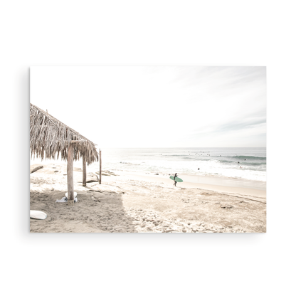 Fine art of WindanSea Beach, La Jolla California horizontal landscape print by Southern California photographer Kim Dybczak of Salt Creek Prints, featuring a bright, slightly desaturated image shows Windansea Beach in La Jolla, California. A thatched beach hut stands on the sandy shore in the left foreground. Further down the beach, a surfer carrying a teal surfboard walks towards the ocean where several other surfers are visible in the water. Photograph in minimalist unframed mock up