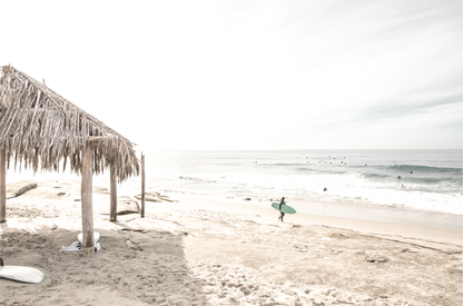 Fine art of WindanSea Beach, La Jolla California horizontal landscape print by Southern California photographer Kim Dybczak of Salt Creek Prints, featuring a bright, slightly desaturated image shows Windansea Beach in La Jolla, California. A thatched beach hut stands on the sandy shore in the left foreground. Further down the beach, a surfer carrying a teal surfboard walks towards the ocean where several other surfers are visible in the water. 