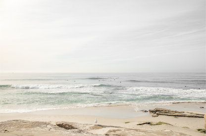 Fine art of horizontal landscape print by Southern California photographer Kim Dybczak of Salt Creek Prints, featuring a bright image captures a pale sandy beach giving way to a vast, light gray ocean with gentle waves. Several surfers are scattered across the water, some sitting on their boards and others riding small waves. The sky is a uniform, pale white, creating a soft, overcast atmosphere, located in Windansea Beach in La Jolla, California.