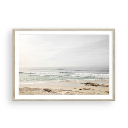 Fine art of horizontal landscape print by Southern California photographer Kim Dybczak of Salt Creek Prints, featuring a bright image captures a pale sandy beach giving way to a vast, light gray ocean with gentle waves. Several surfers are scattered across the water, some sitting on their boards and others riding small waves. The sky is a uniform, pale white, creating a soft, overcast atmosphere, located in Windansea Beach in La Jolla, California. Photograph in framed minimalist mock up