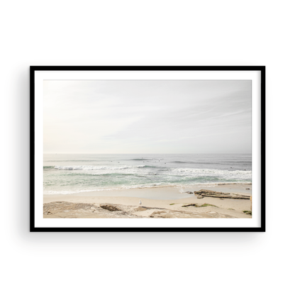 Fine art of horizontal landscape print by Southern California photographer Kim Dybczak of Salt Creek Prints, featuring a bright image captures a pale sandy beach giving way to a vast, light gray ocean with gentle waves. Several surfers are scattered across the water, some sitting on their boards and others riding small waves. The sky is a uniform, pale white, creating a soft, overcast atmosphere, located in Windansea Beach in La Jolla, California. Photograph in framed minimalist mock up