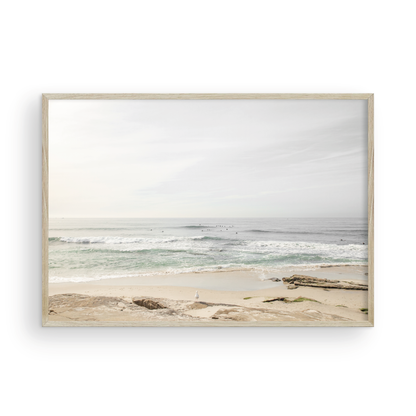 Fine art of horizontal landscape print by Southern California photographer Kim Dybczak of Salt Creek Prints, featuring a bright image captures a pale sandy beach giving way to a vast, light gray ocean with gentle waves. Several surfers are scattered across the water, some sitting on their boards and others riding small waves. The sky is a uniform, pale white, creating a soft, overcast atmosphere, located in Windansea Beach in La Jolla, California. Photograph in framed minimalist mock up
