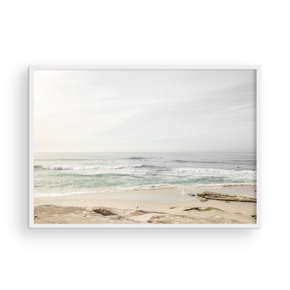 Fine art of horizontal landscape print by Southern California photographer Kim Dybczak of Salt Creek Prints, featuring a bright image captures a pale sandy beach giving way to a vast, light gray ocean with gentle waves. Several surfers are scattered across the water, some sitting on their boards and others riding small waves. The sky is a uniform, pale white, creating a soft, overcast atmosphere, located in Windansea Beach in La Jolla, California. Photograph in framed minimalist mock up