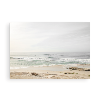 Fine art of horizontal landscape print by Southern California photographer Kim Dybczak of Salt Creek Prints, featuring a bright image captures a pale sandy beach giving way to a vast, light gray ocean with gentle waves. Several surfers are scattered across the water, some sitting on their boards and others riding small waves. The sky is a uniform, pale white, creating a soft, overcast atmosphere, located in Windansea Beach in La Jolla, California. Photograph in unframed minimalist mock up