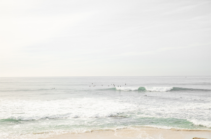 Fine art of horizontal landscape print by Southern California photographer Kim Dybczak of Salt Creek Prints, featuring a bright image captures a pale sandy beach giving way to a vast, light gray ocean with gentle waves. Several surfers are scattered across the water, some sitting on their boards and others riding small waves. The sky is a uniform, pale white, creating a soft, overcast atmosphere, located in Windansea Beach in La Jolla, California. 