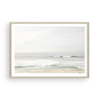 Fine art of horizontal landscape print by Southern California photographer Kim Dybczak of Salt Creek Prints, featuring a bright image captures a pale sandy beach giving way to a vast, light gray ocean with gentle waves. Several surfers are scattered across the water, some sitting on their boards and others riding small waves. The sky is a uniform, pale white, creating a soft, overcast atmosphere, located in Windansea Beach in La Jolla, California. Photograph in framed minimalist mock up
