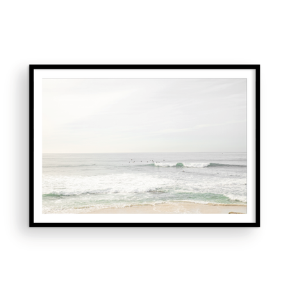 Fine art of horizontal landscape print by Southern California photographer Kim Dybczak of Salt Creek Prints, featuring a bright image captures a pale sandy beach giving way to a vast, light gray ocean with gentle waves. Several surfers are scattered across the water, some sitting on their boards and others riding small waves. The sky is a uniform, pale white, creating a soft, overcast atmosphere, located in Windansea Beach in La Jolla, California. Photograph in framed minimalist mock up