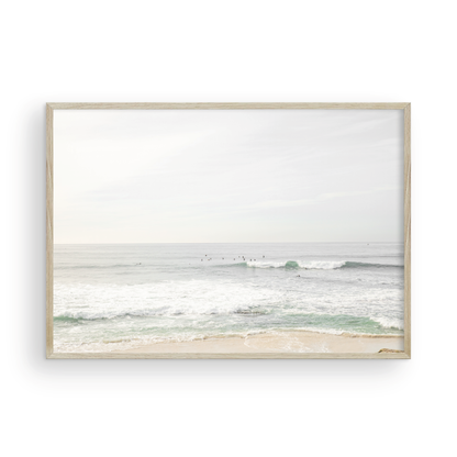Fine art of horizontal landscape print by Southern California photographer Kim Dybczak of Salt Creek Prints, featuring a bright image captures a pale sandy beach giving way to a vast, light gray ocean with gentle waves. Several surfers are scattered across the water, some sitting on their boards and others riding small waves. The sky is a uniform, pale white, creating a soft, overcast atmosphere, located in Windansea Beach in La Jolla, California. Photograph in framed minimalist mock up