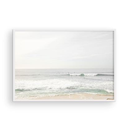 Fine art of horizontal landscape print by Southern California photographer Kim Dybczak of Salt Creek Prints, featuring a bright image captures a pale sandy beach giving way to a vast, light gray ocean with gentle waves. Several surfers are scattered across the water, some sitting on their boards and others riding small waves. The sky is a uniform, pale white, creating a soft, overcast atmosphere, located in Windansea Beach in La Jolla, California. Photograph in framed minimalist mock up