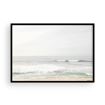 Fine art of horizontal landscape print by Southern California photographer Kim Dybczak of Salt Creek Prints, featuring a bright image captures a pale sandy beach giving way to a vast, light gray ocean with gentle waves. Several surfers are scattered across the water, some sitting on their boards and others riding small waves. The sky is a uniform, pale white, creating a soft, overcast atmosphere, located in Windansea Beach in La Jolla, California. Photograph in framed minimalist mock up