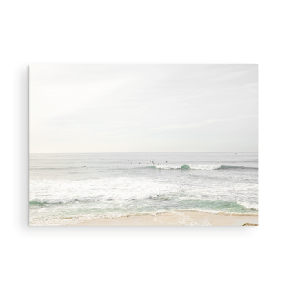 Fine art of horizontal landscape print by Southern California photographer Kim Dybczak of Salt Creek Prints, featuring a bright image captures a pale sandy beach giving way to a vast, light gray ocean with gentle waves. Several surfers are scattered across the water, some sitting on their boards and others riding small waves. The sky is a uniform, pale white, creating a soft, overcast atmosphere, located in Windansea Beach in La Jolla, California. Photograph in unframed minimalist mock up
