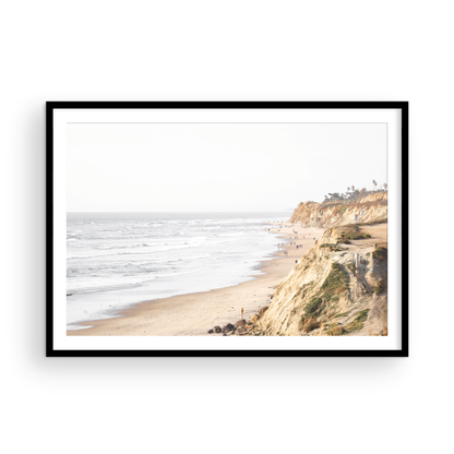 Fine art of Torrey Pines, San Diego California horizontal print of hazy high-angle landscape beach photography captures a light blue ocean meeting a sandy beach with many beach goers. To the right, tall tan and brown cliffs rise as iconic coastal scenery. Photograph in minimalist framed mock up.