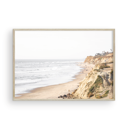 Fine art of Torrey Pines, San Diego California horizontal print of hazy high-angle landscape beach photography captures a light blue ocean meeting a sandy beach with many beach goers. To the right, tall tan and brown cliffs rise as iconic coastal scenery. Photograph in minimalist framed mock up.