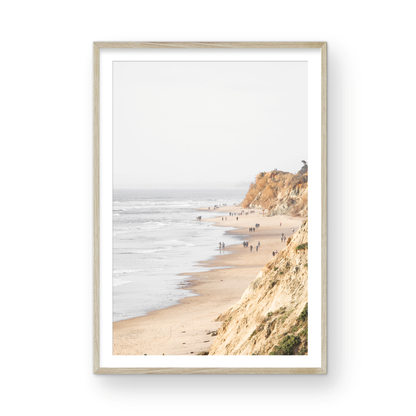 Fine art of Torrey Pines, San Diego California vertical print of hazy high-angle landscape beach photography captures a light blue ocean meeting a sandy beach with many beach goers. To the right, tall tan and brown cliffs rise as iconic coastal scenery. Photograph in minimalist framed mock up.