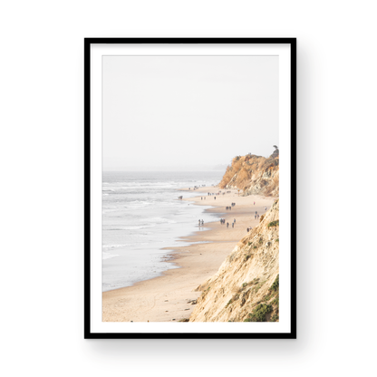 Fine art of Torrey Pines, San Diego California vertical print of hazy high-angle landscape beach photography captures a light blue ocean meeting a sandy beach with many beach goers. To the right, tall tan and brown cliffs rise as iconic coastal scenery. Photograph in minimalist framed mock up.