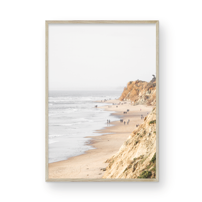 Fine art of Torrey Pines, San Diego California vertical print of hazy high-angle landscape beach photography captures a light blue ocean meeting a sandy beach with many beach goers. To the right, tall tan and brown cliffs rise as iconic coastal scenery. Photograph in minimalist framed mock up.