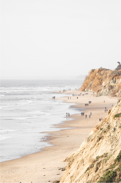 Fine art of Torrey Pines, San Diego California vertical print of hazy high-angle landscape beach photography captures a light blue ocean meeting a sandy beach with many beach goers. To the right, tall tan and brown cliffs rise as iconic coastal scenery. Photograph in minimalist framed mock up.