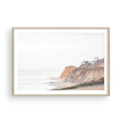 Fine art of Torrey Pines, San Diego California horizontal print of hazy high-angle landscape beach photography captures a light blue ocean meeting a sandy beach with many beach goers. To the right, tall tan and brown cliffs rise as iconic coastal scenery. Photograph in minimalist framed mock up.