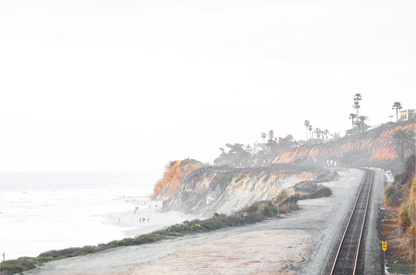 A slightly hazy, wide Landscape Coastal Photography shot shows a set of train tracks curving along a coastal cliffside, San Diego Coast. To the left, a sandy beach meets a light blue ocean waves with gentle waves, where a few small figures of people are visible. The cliffs are varied in color, with sections of tan, white, and hints of orange, topped with greenery and a few Palm Trees. The sky is a muted white, perfect for Minimalist Surf Wall Art.