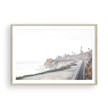 A slightly hazy, wide Landscape Coastal Photography shot shows a set of train tracks curving along a coastal cliffside, San Diego Coast. To the left, a sandy beach meets a light blue ocean waves with gentle waves, where a few small figures of people are visible. The cliffs are varied in color, with sections of tan, white, and hints of orange, topped with greenery and a few Palm Trees. The sky is a muted white, perfect for Minimalist Surf Wall Art. Photograph in minimalist mock up