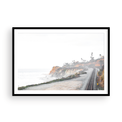 A slightly hazy, wide Landscape Coastal Photography shot shows a set of train tracks curving along a coastal cliffside, San Diego Coast. To the left, a sandy beach meets a light blue ocean waves with gentle waves, where a few small figures of people are visible. The cliffs are varied in color, with sections of tan, white, and hints of orange, topped with greenery and a few Palm Trees. The sky is a muted white, perfect for Minimalist Surf Wall Art. Photograph in minimalist mock up