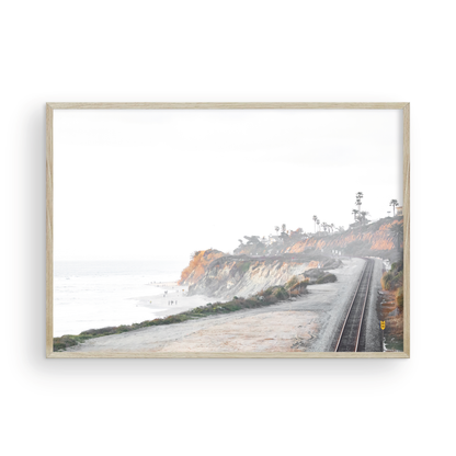 A slightly hazy, wide Landscape Coastal Photography shot shows a set of train tracks curving along a coastal cliffside, San Diego Coast. To the left, a sandy beach meets a light blue ocean waves with gentle waves, where a few small figures of people are visible. The cliffs are varied in color, with sections of tan, white, and hints of orange, topped with greenery and a few Palm Trees. The sky is a muted white, perfect for Minimalist Surf Wall Art. Photograph in minimalist mock up