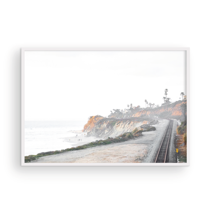 A slightly hazy, wide Landscape Coastal Photography shot shows a set of train tracks curving along a coastal cliffside, San Diego Coast. To the left, a sandy beach meets a light blue ocean waves with gentle waves, where a few small figures of people are visible. The cliffs are varied in color, with sections of tan, white, and hints of orange, topped with greenery and a few Palm Trees. The sky is a muted white, perfect for Minimalist Surf Wall Art. Photograph in minimalist mock up