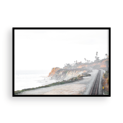 A slightly hazy, wide Landscape Coastal Photography shot shows a set of train tracks curving along a coastal cliffside, San Diego Coast. To the left, a sandy beach meets a light blue ocean waves with gentle waves, where a few small figures of people are visible. The cliffs are varied in color, with sections of tan, white, and hints of orange, topped with greenery and a few Palm Trees. The sky is a muted white, perfect for Minimalist Surf Wall Art. Photograph in minimalist mock up