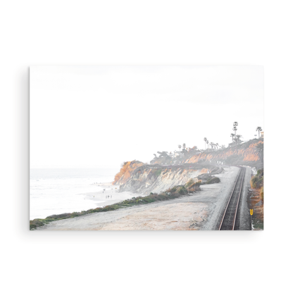 A slightly hazy, wide Landscape Coastal Photography shot shows a set of train tracks curving along a coastal cliffside, San Diego Coast. To the left, a sandy beach meets a light blue ocean waves with gentle waves, where a few small figures of people are visible. The cliffs are varied in color, with sections of tan, white, and hints of orange, topped with greenery and a few Palm Trees. The sky is a muted white, perfect for Minimalist Surf Wall Art. Photograph in minimalist mock up