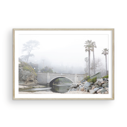 Landscape coastal photography of a concrete arch bridge disappearing into a dense fog, flanked by palm trees and rocky shores. This calm beach print evokes a mysterious seascape. Photograph in minimalist mock up.